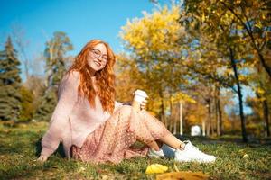 ritratti di un'affascinante ragazza dai capelli rossi con occhiali e un bel viso. ragazza in posa nel parco autunnale con un maglione e una gonna color corallo. foto