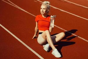 giovane bella bionda si siede su una pista da jogging con una bottiglia d'acqua in mano foto