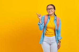 sorridente giovane studentessa asiatica in abito denim con zaino che punta il dito nello spazio della copia, mostrando prodotti pubblicitari isolati su sfondo giallo. istruzione nel concetto di college universitario foto