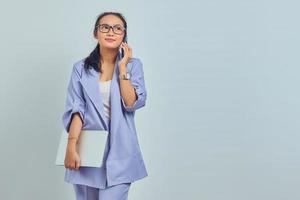 Ritratto di giovane donna asiatica sorridente che parla sul telefono cellulare e tiene il laptop, in piedi guardando lateralmente isolato su sfondo bianco foto