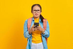 ritratto di giovane studentessa asiatica infastidita in abiti casual con zaino utilizzando il telefono cellulare e guardando la fotocamera isolata su sfondo giallo. istruzione nel concetto di università universitaria foto