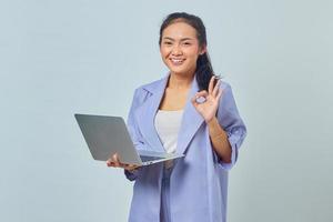 Ritratto di giovane donna asiatica sorridente con laptop e gesticolando segno ok isolato su sfondo bianco foto