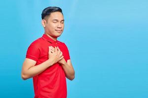 ritratto di bel giovane asiatico in camicia rossa premendo le mani sul petto con gli occhi chiusi e gesto grato sul viso. concetto di salute. foto