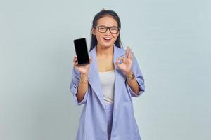 Ritratto di giovane donna asiatica sorridente che mostra il telefono cellulare con schermo vuoto e che gesturing il segno giusto isolato su priorità bassa bianca foto