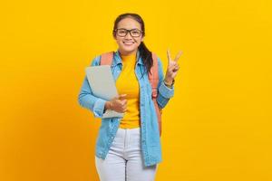 Ritratto di giovane studentessa asiatica allegra in abiti casual con zaino in possesso di laptop e che mostra il segno di pace con il dito isolato su sfondo giallo. istruzione nel concetto di college universitario foto