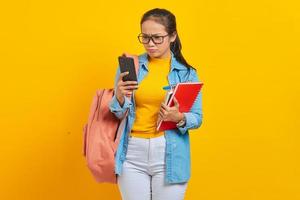 Ritratto di giovane studentessa asiatica seria in abiti denim con zaino utilizzando il telefono cellulare e tenere libri isolati su sfondo giallo foto