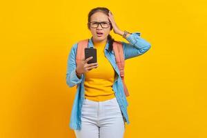 ritratto di giovane studentessa asiatica sorpresa in abiti casual con zaino utilizzando il telefono cellulare con la bocca aperta e tenendo le mani in testa isolato su sfondo giallo foto