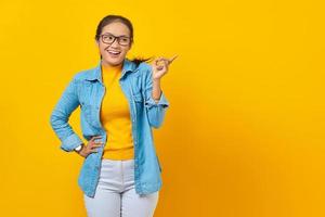 Ritratto di giovane studentessa asiatica sorridente in abiti denim che punta il dito nello spazio della copia, mostrando prodotti pubblicitari isolati su sfondo giallo. istruzione nel concetto di università universitaria foto