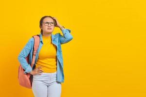 bella giovane studentessa asiatica in abito denim con zaino e guardando seriamente pensando alla domanda, pensando con la mano sulla testa su sfondo giallo. concetto di college universitario di scuola superiore foto