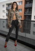 splendida donna bruna glamour con giacca a quadri in posa sul balcone moderno con splendida vista sulla città. ritratto di una donna alla moda alla moda con gambe lunghe, jeans neri che indossano in balcone foto