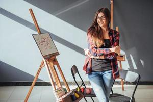 artista ragazza piuttosto carina dipinge su tela dipinto sul cavalletto. sfondo nero da studio. capelli lunghi, bruna. foto