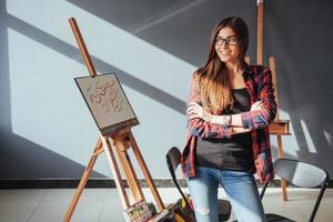 artista ragazza piuttosto carina dipinge su tela dipinto sul cavalletto. sfondo nero da studio. capelli lunghi, bruna. foto