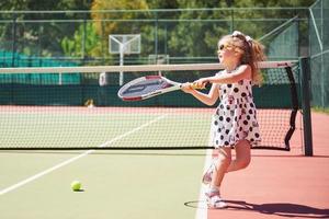 bambina sveglia che gioca a tennis sul campo da tennis all'esterno foto