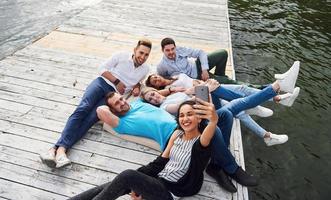 un gruppo di giovani seduti sul bordo del molo, e fanno selfie. amici che si divertono a giocare sul lago foto
