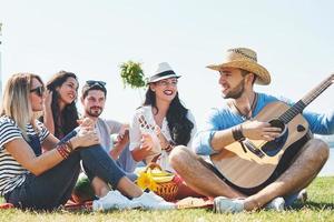 giovani amici felici che fanno un picnic nel parco. sono tutti felici, si divertono, sorridono e suonano la chitarra foto