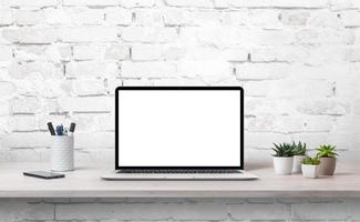 laptop sulla scrivania dell'ufficio con schermo isolato per mockup. piante, telefono e penne accanto. muro di mattoni bianchi sullo sfondo foto