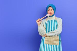 bella donna asiatica pensosa in maglione bianco ricoperta di grembiule e hijab che tiene un cucchiaio, guardando in alto facendo i lavori domestici isolati su sfondo viola. concetto di pulizia foto