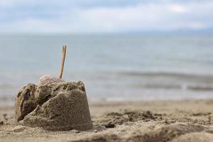 montagna di sabbia con un bastone di legno sulla spiaggia. castello di sabbia con un sasso sulla spiaggia. primo piano di piccoli castelli di sabbia con un bastone sormontato da un cielo blu foto