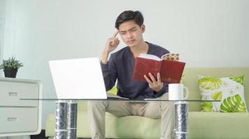 uomo asiatico che tiene libro mentre pensa allo schermo del laptop a casa foto