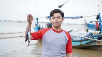 felice giovane pescatore sulla spiaggia che tiene il suo pesce pescato e mostra davanti alla sua barca foto