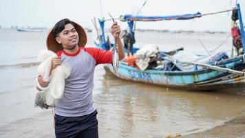 felice giovane pescatore sulla spiaggia che tiene il suo pesce pescato e mostra davanti alla sua barca foto