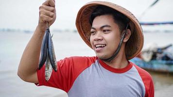 felice giovane pescatore sulla spiaggia che tiene il suo pesce pescato e mostra davanti alla sua barca foto