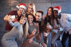 raggruppare bei giovani che fanno selfie nella festa di capodanno, migliori amici ragazze e ragazzi che si divertono insieme, posando persone con uno stile di vita emotivo. cappelli di Babbo Natale e bicchieri di champagne nelle loro mani