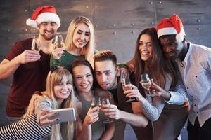gruppo di bei giovani che fanno selfie nella festa di capodanno, migliori amici ragazze e ragazzi insieme divertendosi, in posa concetto di persone di stile di vita. cappelli di Babbo Natale e bicchieri di champagne nelle loro mani foto
