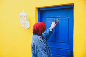 donna musulmana che bussa alla porta di legno blu sulla parete gialla della casa foto