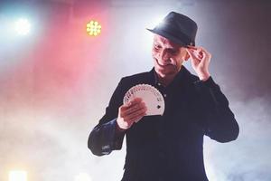 mago che mostra trucco con le carte da gioco. magia o destrezza, circo, gioco d'azzardo. prestigiatore in camera oscura con nebbia foto