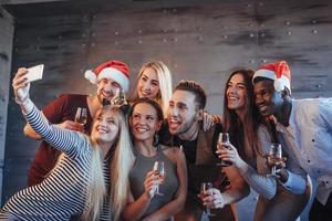 raggruppare bei giovani che fanno selfie nella festa di capodanno, migliori amici ragazze e ragazzi che si divertono insieme, posando persone con uno stile di vita emotivo. cappelli di Babbo Natale e bicchieri di champagne nelle loro mani foto