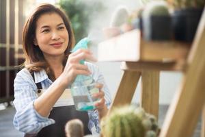 adulto asiatico carino 45s donna che indossa il grembiule che tiene la brocca dello spruzzatore e spruzza goccioline d'acqua al cactus nello scaffale con la faccia sorridente felice a casa. tempo di relax e concetto di hobby foto