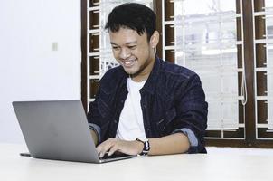 giovane uomo asiatico che si sente felice e sorride quando lavora al computer portatile sul tavolo. uomo indonesiano che indossa una camicia blu. foto