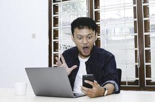 il giovane asiatico è scioccato e stupisce quello che vede nel laptop quando è in chiamata sullo smartphone foto
