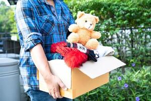 tenendo in mano una scatola per la donazione di vestiti con vestiti usati e una bambola a casa per sostenere l'aiuto ai poveri nel mondo. foto