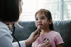 la bambina asiatica sta sorridendo e si siede sul divano a casa durante una visita medica mentre la professionista medica femminile sta usando uno stetoscopio per ascoltare il suo battito cardiaco. foto