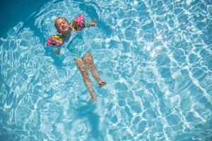 carina bambina che nuota in piscina turchese con braccia gonfiabili aiuta il supporto, vista dall'alto. foto