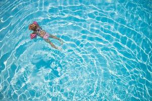 carina bambina che nuota in piscina turchese con braccia gonfiabili aiuta il supporto, vista dall'alto. foto