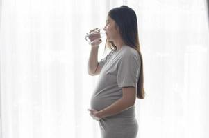 giovane donna incinta che beve acqua a casa, assistenza sanitaria e concetto di cura della gravidanza foto