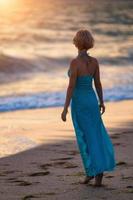 una giovane ragazza snella si erge sulla spiaggia al tramonto con un abito lungo blu, una bella figura foto