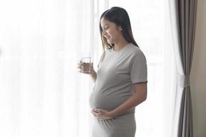 giovane donna incinta che beve acqua a casa, assistenza sanitaria e concetto di cura della gravidanza foto