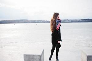 ragazza casual al cappotto nero, sciarpa e cappello contro il fiume ghiacciato in un clima invernale soleggiato. foto