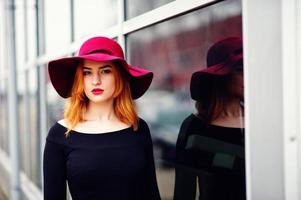 ritratto di moda ragazza dai capelli rossi su cappello rosso e vestito nero con trucco luminoso poste contro la grande finestra. filtri instagram in stile foto dai toni.