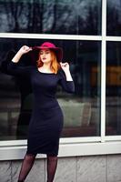 ritratto di moda ragazza dai capelli rossi su cappello rosso e vestito nero con trucco luminoso poste contro la grande finestra. filtri instagram in stile foto dai toni.