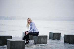 elegante e ricca ragazza bionda su camicetta e pantaloni di pelle con smartphone rosa a portata di mano cubetti di pietra contro il lago ghiacciato il giorno d'inverno. foto