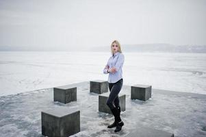 elegante e ricca ragazza bionda su camicetta e pantaloni di pelle con smartphone rosa a portata di mano cubetti di pietra contro il lago ghiacciato il giorno d'inverno. foto