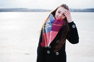 ragazza casual al cappotto nero, sciarpa e cappello contro il fiume ghiacciato in un clima invernale soleggiato. foto