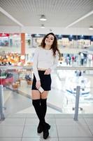 giovane modella riccia con il telefono a portata di mano posata su minigonna in un grande centro commerciale vicino a ringhiere di vetro. foto