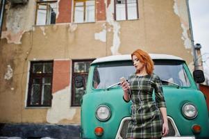giovane ragazza dai capelli rossi signora con cellulare e cuffie, indossando su sfondo vestito controllato vecchio bus minivan turchese vintage. foto