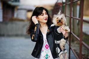 ragazza gitana bruna con cane yorkshire terrier posato contro ringhiere in acciaio. indossa il modello su giacca di pelle e t-shirt con ornamento, pantaloni. foto